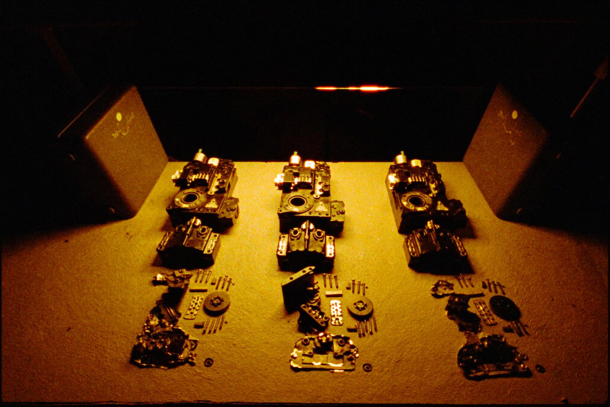 A grainy amber photograph of a dark workshop bench under one hard lamp: three identical machines lie side by side, each stripped completely down to its parts, every plate and screw and fastener laid out in neat rows beneath its own emptied carcass.