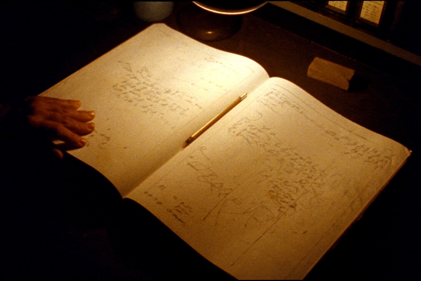 A 35mm film still in tungsten light: an open ledger covered in faint, illegible pencil script, a pencil lying in the gutter, a block eraser waiting at the edge of the light, and a hand resting on the page.