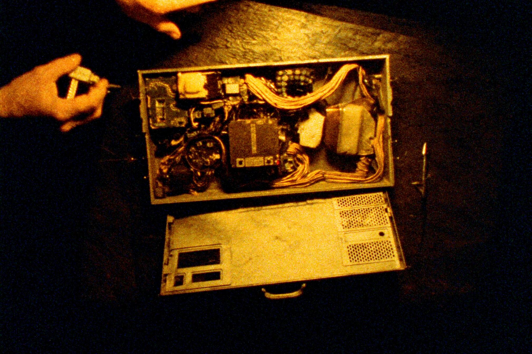 A grainy amber photograph shot from above: a small machine lies open on a dark surface with its cover laid off below it, the boards and ribbon cables inside fully exposed, while a pair of hands enters the frame from the left holding a small tool.