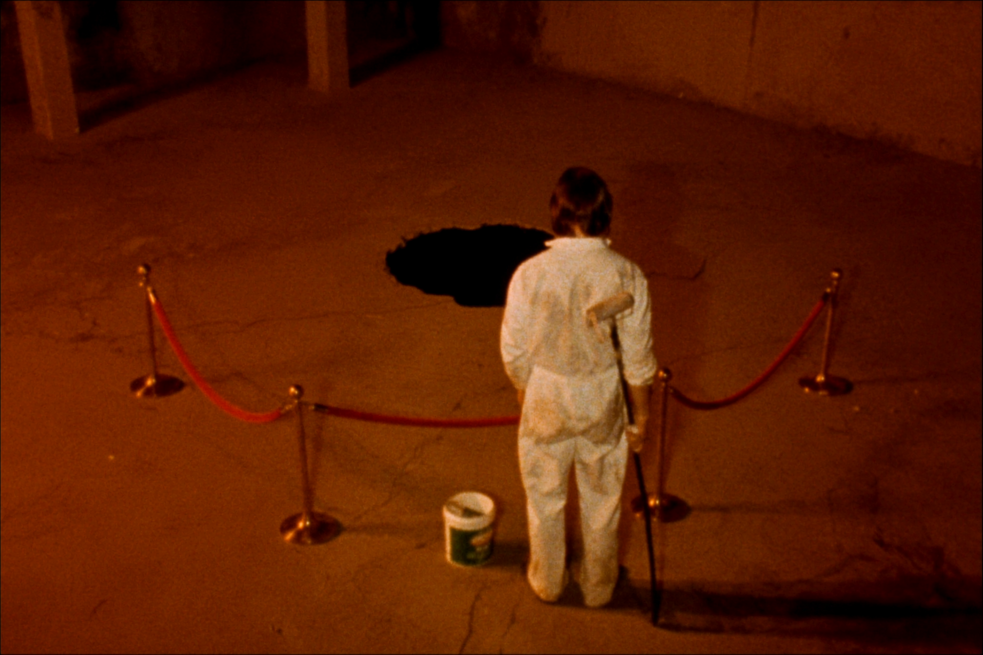 A grainy, sodium-orange photograph of a person in white coveralls standing behind velvet ropes and looking into a perfectly black hole in the floor. A paint roller is tucked behind their back and a bucket sits nearby.